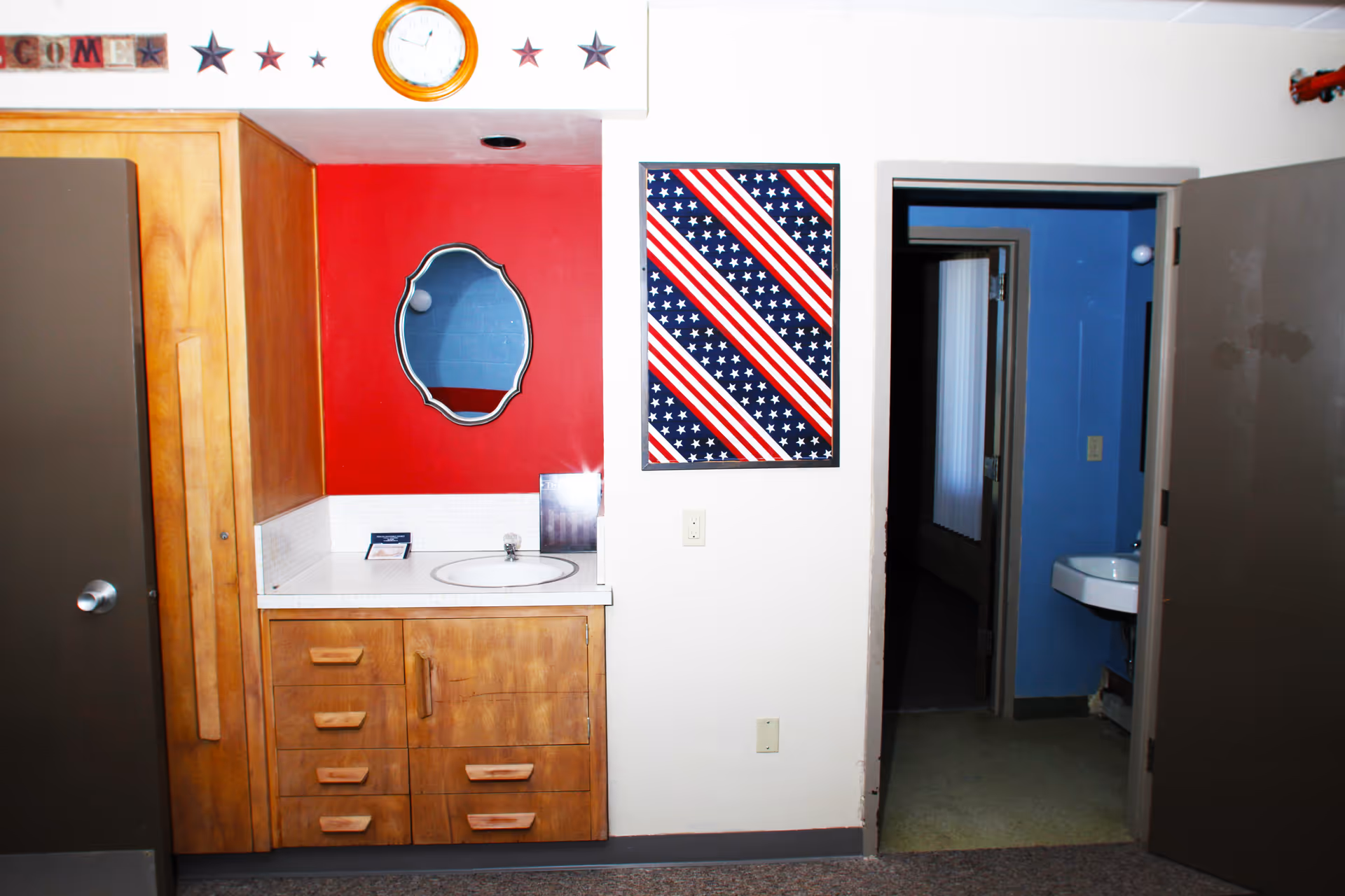 Interior view of a room with a wooden cabinet and sink on the left side, a red wall with a decorative mirror above the sink, and a clock and star decorations above. To the right, there is a doorway leading to another room with a blue wall and a white sink visible.
