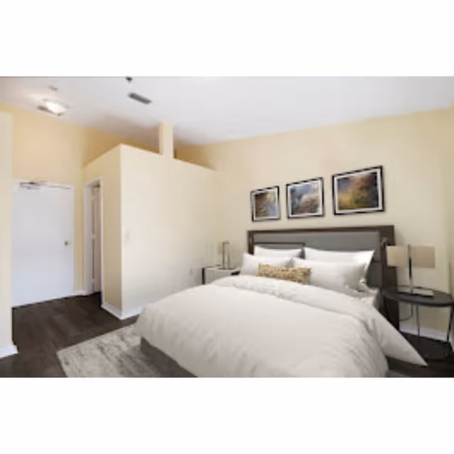 Bright furnished bedroom with a made bed, nightstands, framed art above the headboard, and a doorway to a hallway.