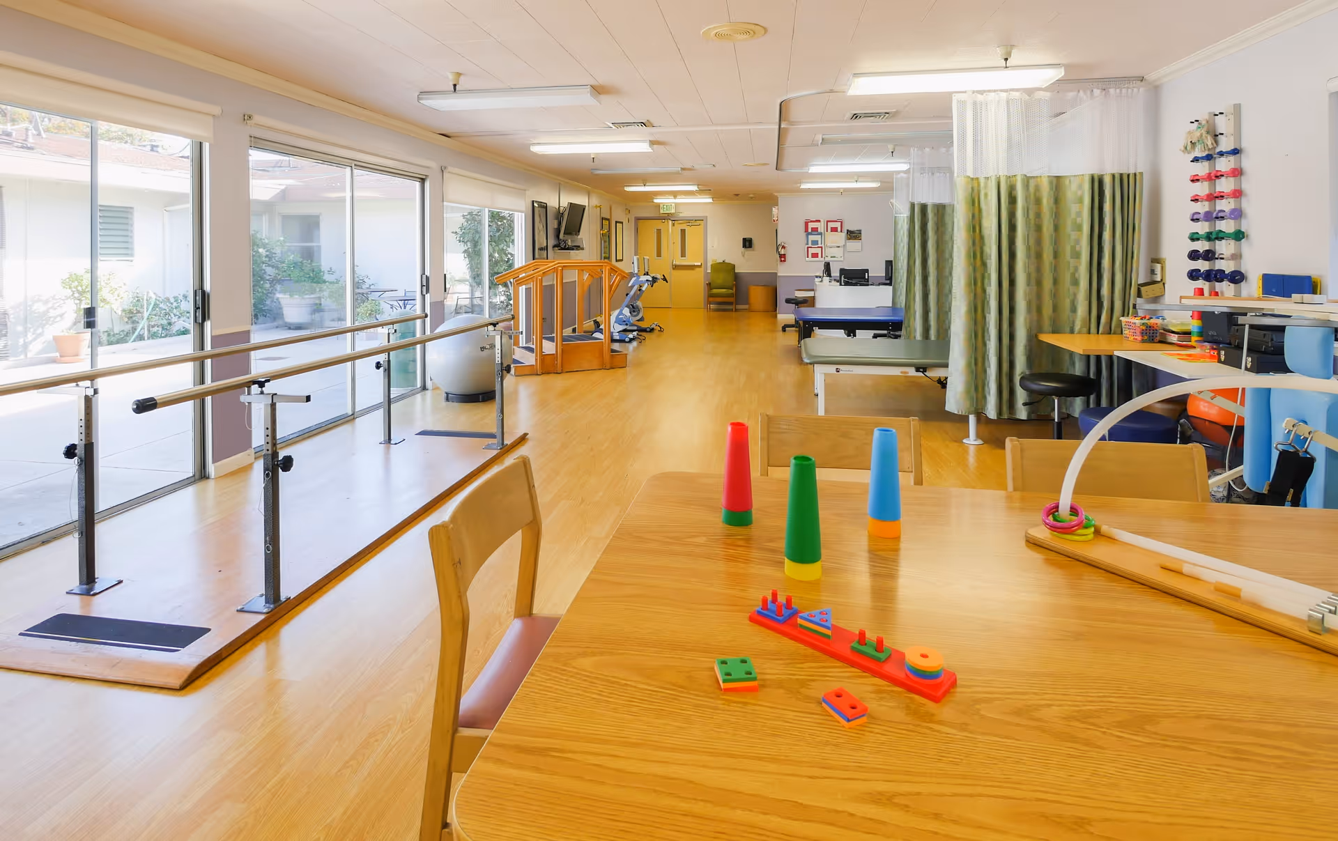 A bright physical therapy room with wooden flooring and large windows letting in natural light. The room contains parallel bars for walking exercises, exercise equipment, therapy tables with privacy curtains, and a wooden table with colorful therapy toys and chairs around it.