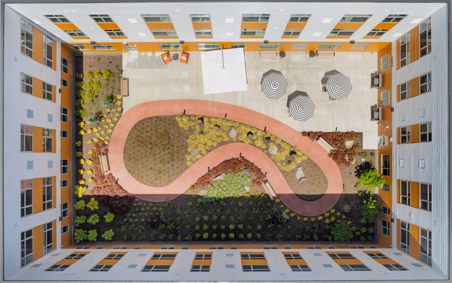 Aerial view of an enclosed courtyard at Clearwater Beaverton facility featuring a winding red pathway surrounded by landscaped garden beds with various plants and shrubs. The courtyard includes several benches, a large white umbrella, and three black and white striped umbrellas providing shade over seating areas. The courtyard is bordered by a multi-story building with white and orange exterior walls and numerous windows.