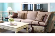 A cozy living room with a beige sofa adorned with multiple cushions, a patterned armchair, a white coffee table with a small flower arrangement and magazines, a floor lamp, and a large window in the background.