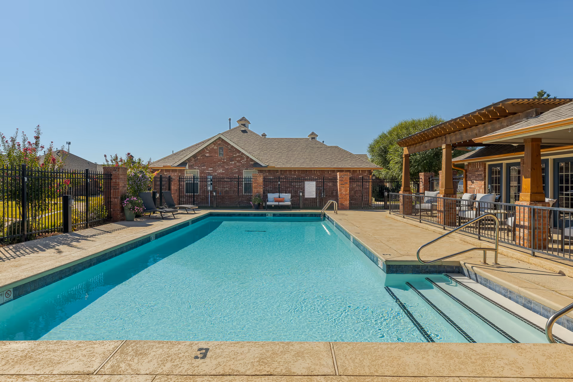 Outdoor swimming pool with clear blue water, surrounded by a concrete deck. There are lounge chairs and potted plants along a black metal fence on the left side. On the right side, there is a covered patio area with wooden beams and seating. In the background, there are brick buildings under a clear blue sky.