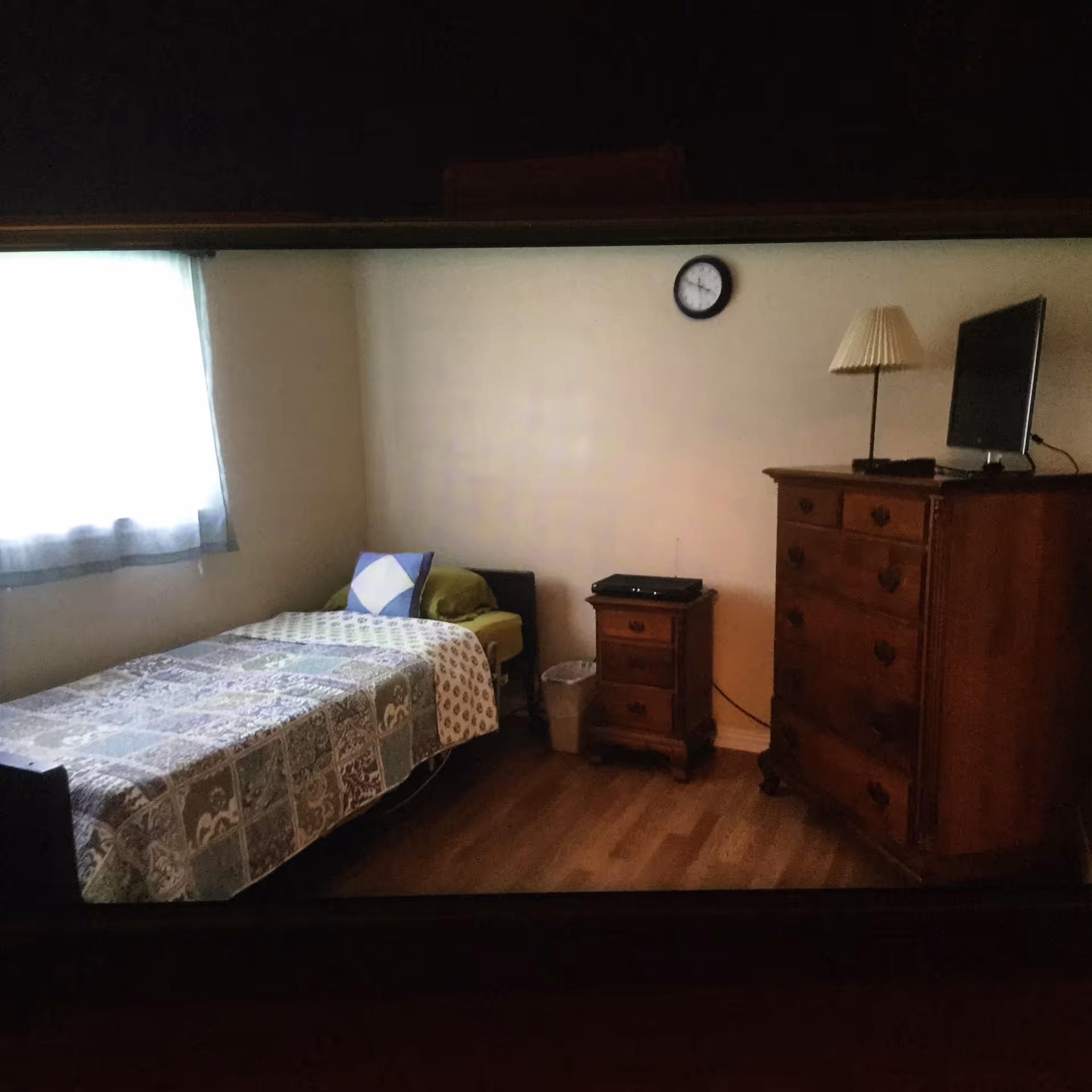 A simple bedroom with a single bed, a bedside table, a wooden dresser with a TV and lamp, a wall clock, and a window.