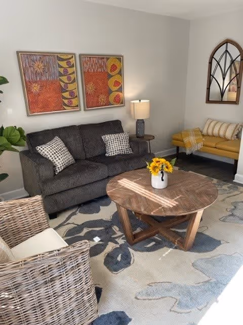 A cozy living room with a dark gray sofa adorned with two patterned pillows, a round wooden coffee table with a vase of yellow flowers, a wicker chair with a white cushion, a yellow bench with a plaid throw, a table lamp on a small side table, two colorful framed artworks on the wall, and a decorative arched mirror.