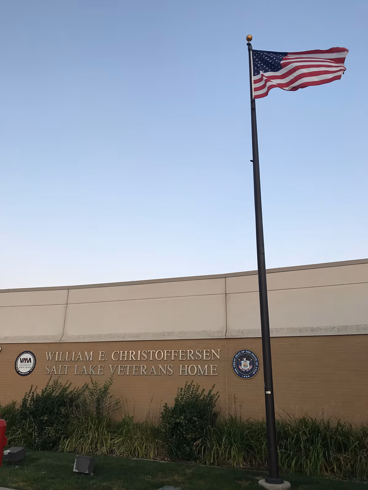Exterior view of the William E. Christoffersen Salt Lake Veterans Home building with an American flag on a tall flagpole in front of it, clear sky in the background, and some bushes and grass near the building.