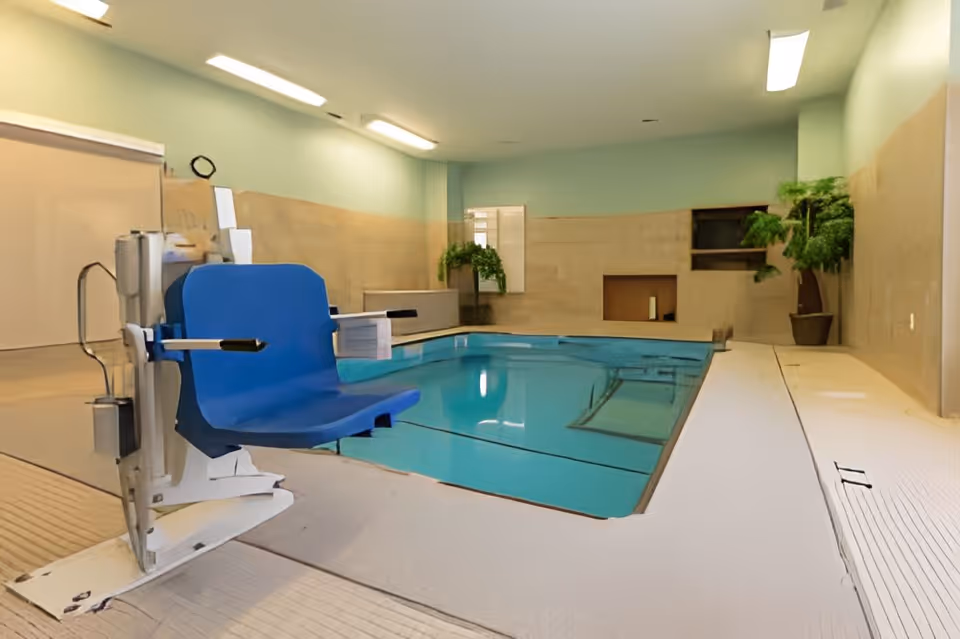 Indoor swimming pool with a blue accessibility lift beside the water and potted plants along the far wall.