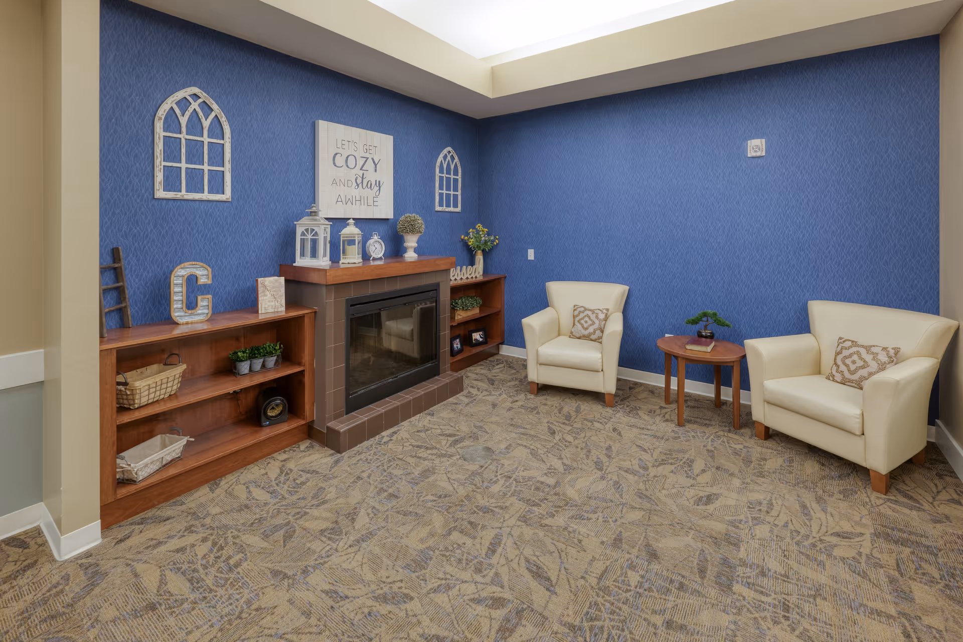 A cozy sitting area with two cream-colored armchairs, a small wooden side table with a plant and a book, a fireplace with a wooden mantel decorated with lanterns, a clock, and a small plant. The walls are painted blue with decorative window frame accents and a sign that reads 'LET'S GET COZY AND stay AWHILE'. The floor is carpeted with a leaf pattern.