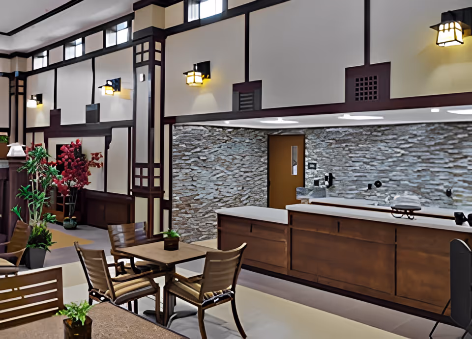 Interior view of a common area in Village Assisted Living featuring wooden tables and chairs, decorative plants, a stone accent wall, and a wooden reception or service counter with warm wall sconces and high windows near the ceiling.