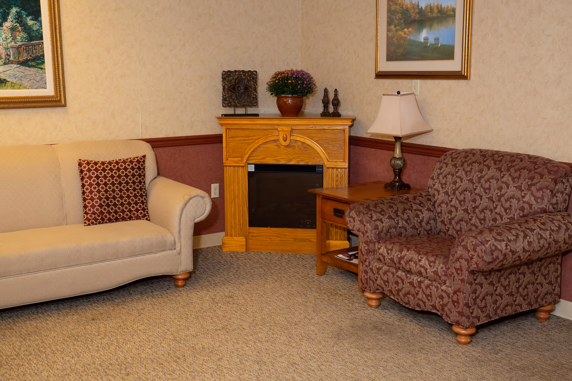 A cozy sitting area with a beige sofa featuring a patterned red cushion on the left, and a patterned maroon armchair on the right. Between them is a wooden electric fireplace with decorative items and a potted plant on top. A wooden side table with a lamp is next to the armchair. The walls are decorated with framed landscape paintings.