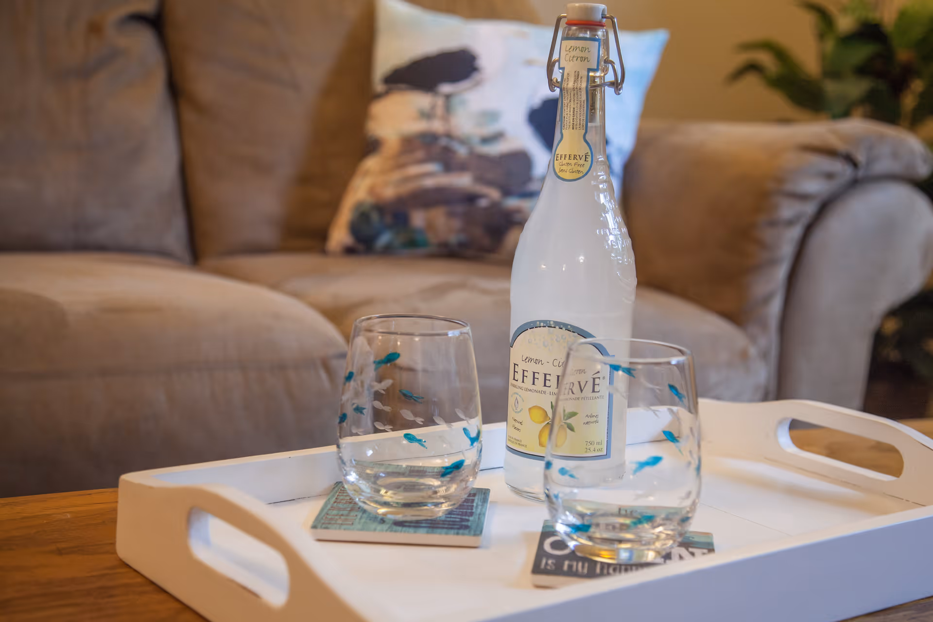 A white tray on a wooden table holding a bottle of lemon citron Effervé sparkling water and two glasses with blue fish designs, placed in front of a beige couch with a decorative pillow.