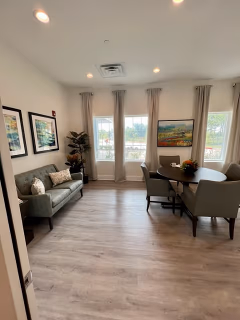 Sunlit communal living area with a sofa, round dining table and chairs by large windows and framed artwork.