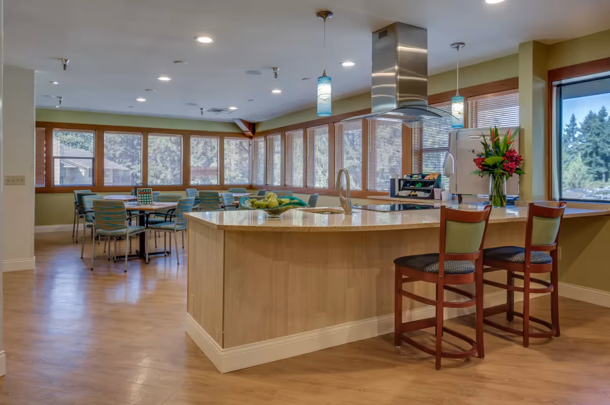 A bright and spacious dining area with a large kitchen island in the foreground. The island has a light-colored countertop with a sink and two wooden chairs with green cushions. Behind the island, there are multiple tables and chairs arranged near large windows with blinds, allowing natural light to fill the room. The walls are painted light green, and there are pendant lights hanging above the island. A vase with flowers and a bowl of fruit are placed on the countertop.