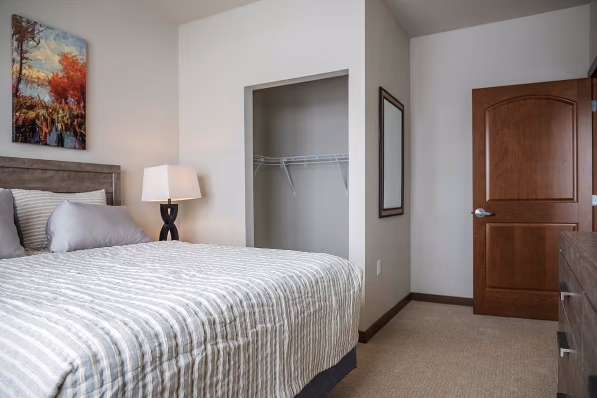 A bedroom with a bed covered in a striped gray and white comforter, two pillows, a wooden headboard, a bedside lamp with a white shade, an open closet with wire shelving, a wooden door, a dresser, a wall mirror, and a colorful painting of trees on the wall.