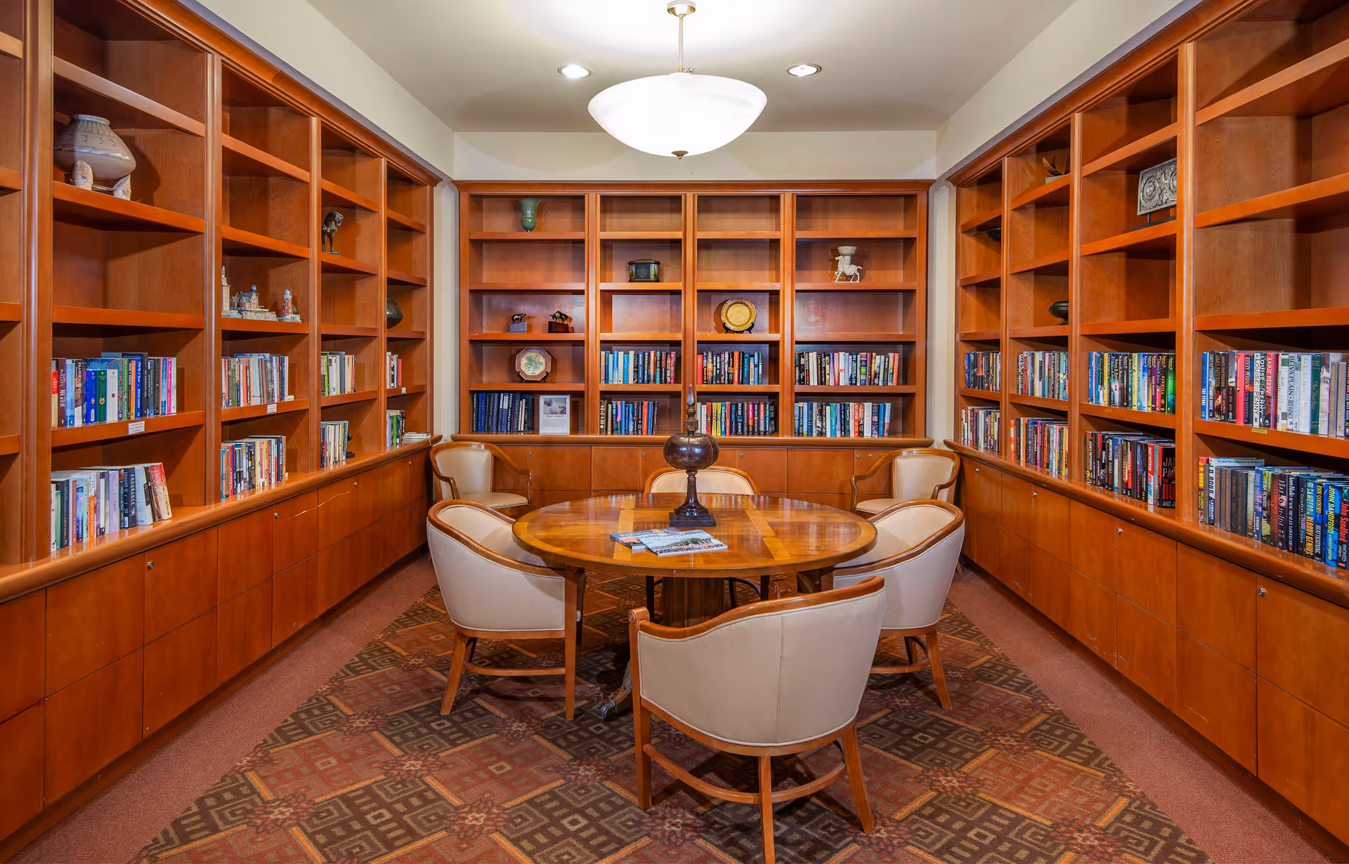 A cozy library/reading room with built-in wooden bookshelves surrounding a round table and six upholstered chairs.