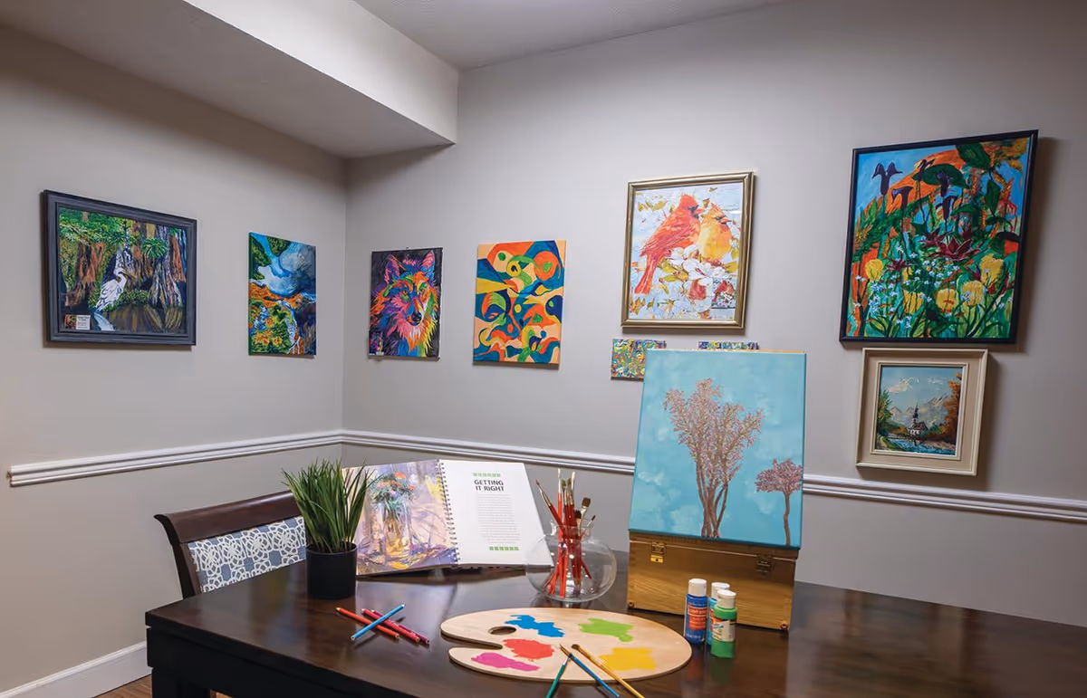 A cozy art corner in a room with a dark wooden table displaying painting supplies including paintbrushes, a palette with colorful paint, and paint bottles. An easel holds a painting of two trees against a blue sky. The wall behind the table is decorated with several colorful framed paintings featuring nature and animals.