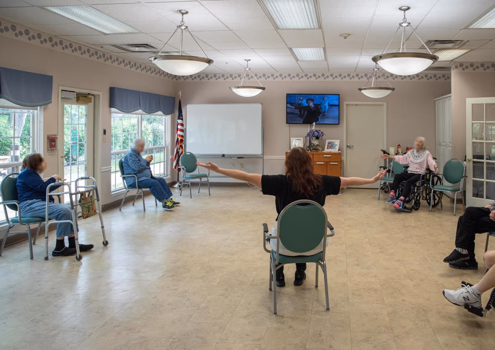 A group of elderly individuals seated in a spacious room arranged in a circle, participating in a seated exercise or stretching session led by an instructor with arms extended. The room has large windows with blue valances, a whiteboard, an American flag, and a television mounted on the wall.