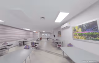 Bright communal dining room with several tables and chairs, wall art, and overhead lighting.