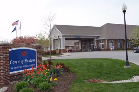 Front exterior of Coventry Meadows senior living facility with entrance, landscaped lawn, and a sign in the foreground.
