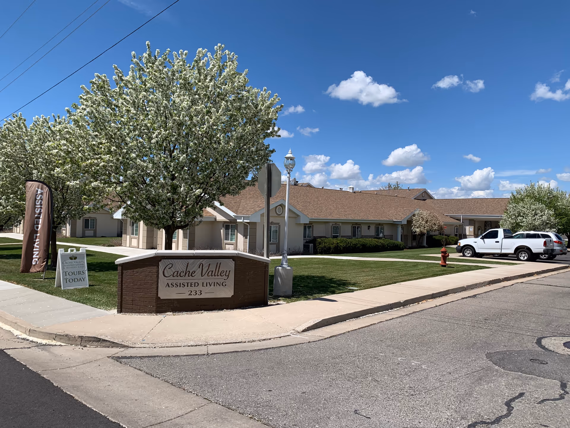 Front entrance sign for Cache Valley Assisted Living in front of a single-story facility with flowering trees and parked vehicles under a blue sky.