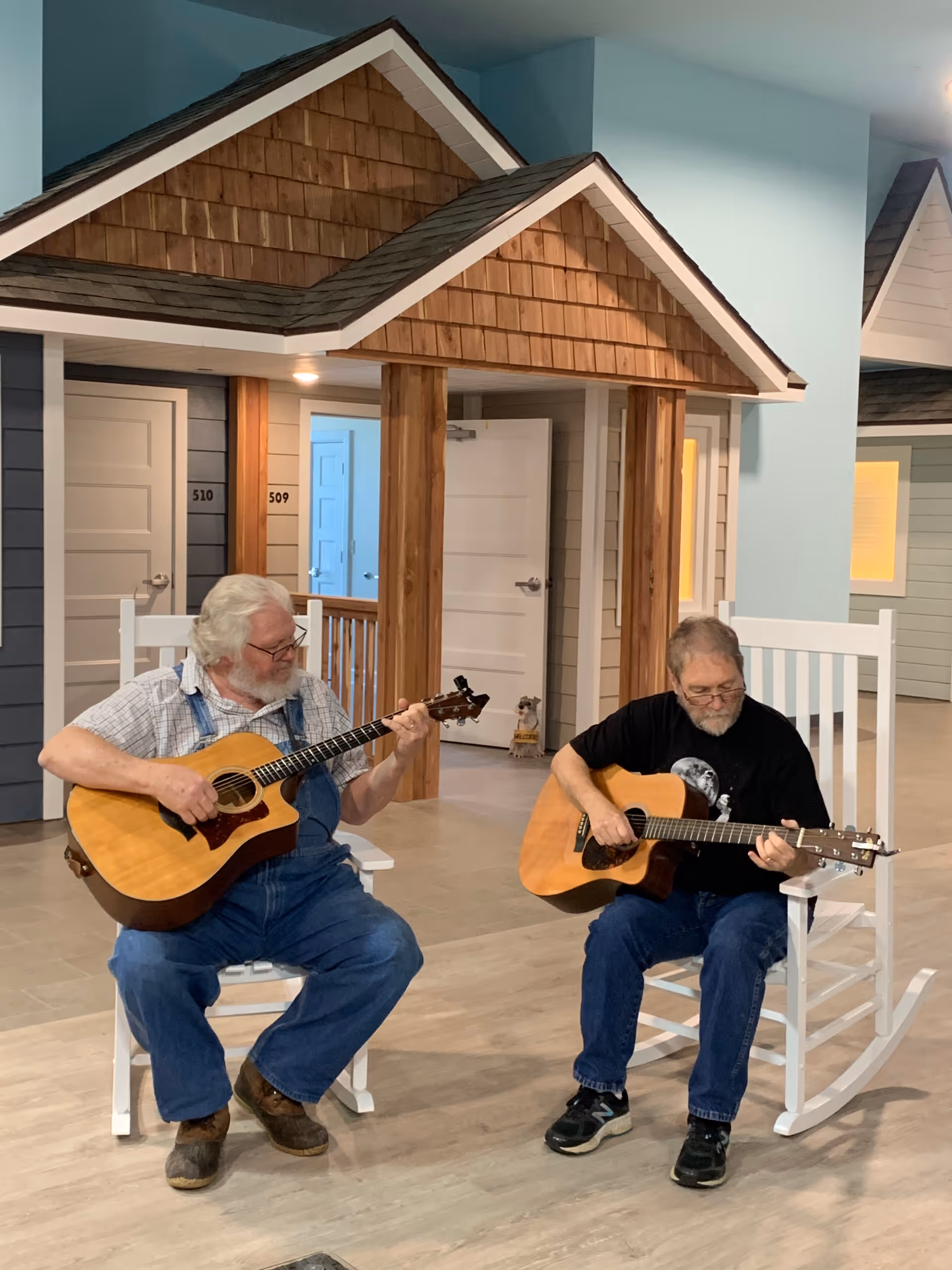 Two elderly men sitting on white rocking chairs indoors, each playing an acoustic guitar. Behind them are small house-like structures with doors numbered 509 and 510, and wooden pillars supporting small peaked roofs.