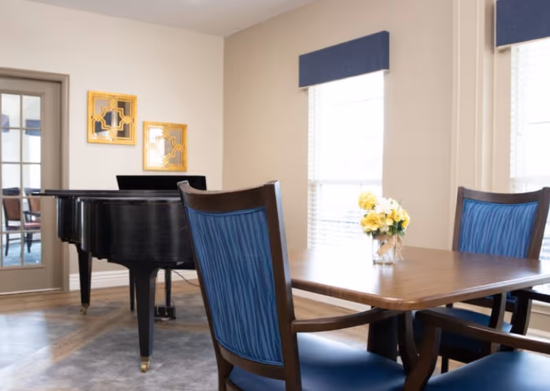 A bright room with a wooden table surrounded by blue cushioned chairs. A small vase with yellow flowers is placed on the table. In the background, there is a black grand piano near a door with glass panels. Two decorative gold-framed mirrors hang on the wall above the piano. The room has large windows with white blinds and blue valances.