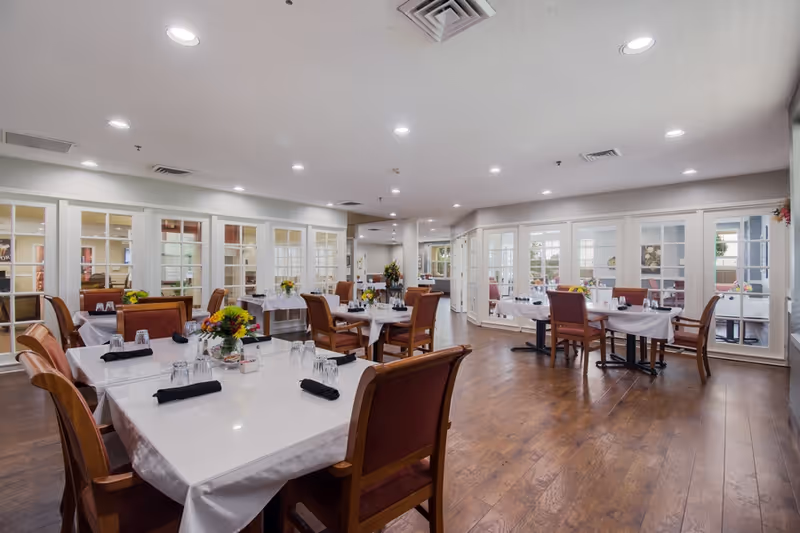 A spacious dining room with multiple tables covered in white tablecloths, each set with glasses and black napkins. Wooden chairs with red cushions surround the tables. The room has wooden flooring, white walls, and large windows with white frames allowing natural light to enter. There are flower arrangements on the tables and ceiling lights providing additional illumination.