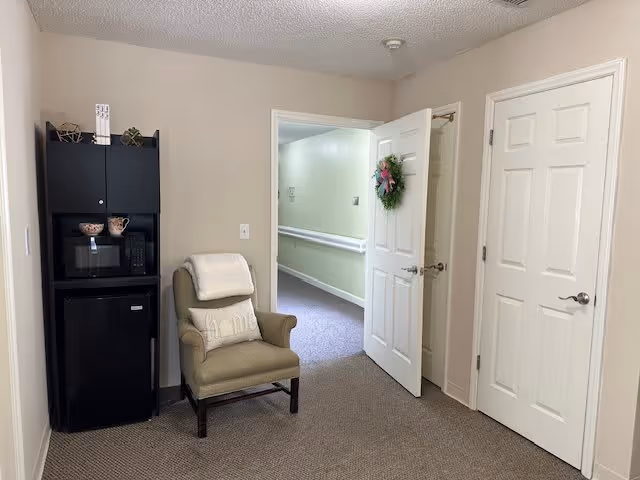 A small interior room with beige walls and carpeted floor featuring a cushioned armchair with a pillow and blanket, a black cabinet with a microwave and mini fridge, and two white doors, one of which is open showing a hallway with handrails and light green walls.