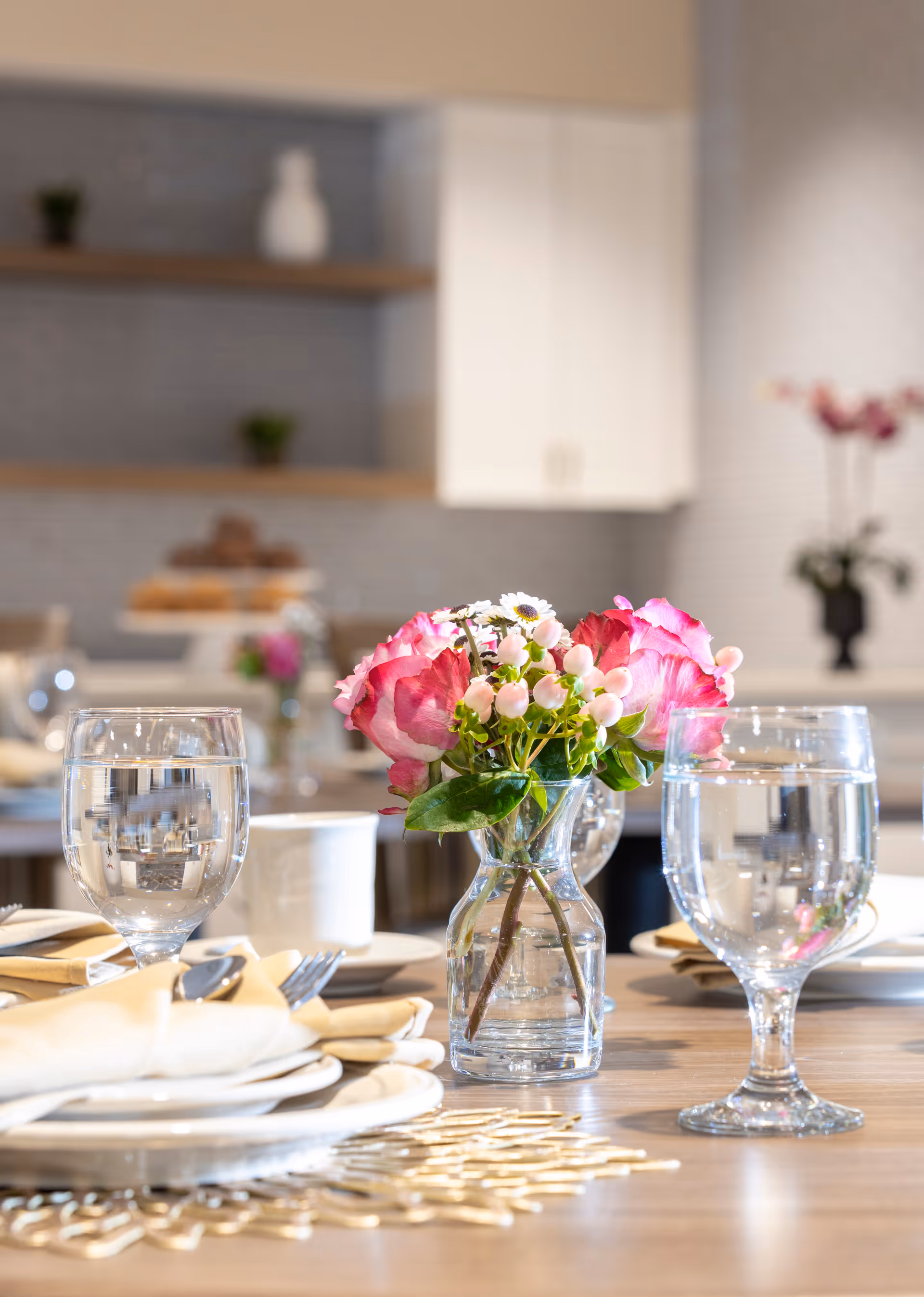 Close-up of a dining table set with plates, beige napkins, silverware, two glasses of water, and a small glass vase with pink and white flowers. The background shows a blurred kitchen area with shelves and cabinets.