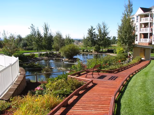 Wooden boardwalk with a bench beside a landscaped pond, trees, and a multi-story building in the background.