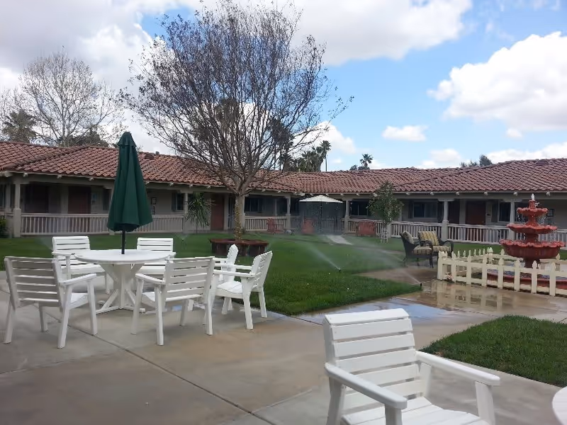Outdoor courtyard area with white patio chairs and tables, a green umbrella, a red multi-tiered fountain, green grass with sprinklers on, and a building with a red tile roof in the background under a partly cloudy sky.