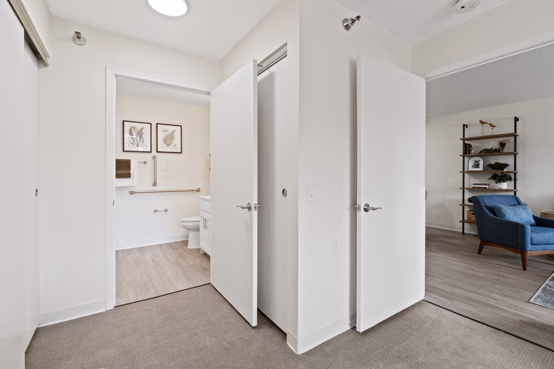 Interior hallway showing an open bathroom with grab bars on the left and a living area with a blue chair and shelving on the right.