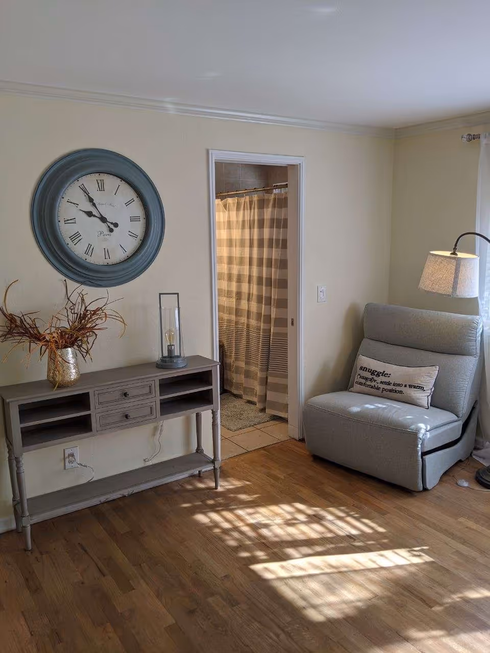 A cozy interior room with wooden flooring featuring a gray armchair with a pillow that reads 'snuggle: to wrap, settle into a warm, comfortable position.' Next to the chair is a floor lamp with a beige shade. On the left wall, there is a gray console table with drawers, decorated with a vase holding dried branches and a glass candle holder. Above the table hangs a large round clock with Roman numerals. An open doorway reveals a bathroom with a striped shower curtain.
