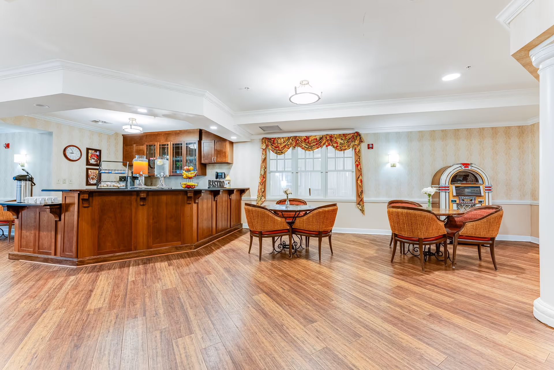 A bright and spacious dining area in a senior living facility featuring a wooden buffet counter with glass cabinets, beverage dispensers, and a fruit stand. There are two round tables with four cushioned chairs each, a window with decorative curtains, and a vintage jukebox against the wall. The room has wood flooring and soft lighting.