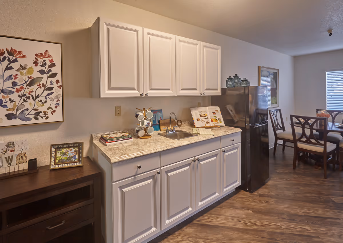A kitchen area with white cabinets, a marble countertop, a small sink, and a black refrigerator. There are decorative items on the countertop including a mug holder, books, and a cookbook. To the right, there is a dining table with chairs near a window with blinds. On the left wall, there is a framed floral artwork and a small wooden side table with picture frames and decorative items.