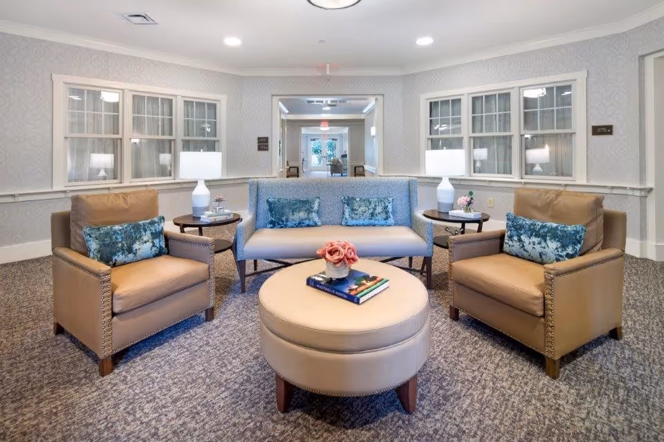 A cozy seating area in a senior living facility with two tan armchairs and a blue loveseat, each adorned with blue patterned throw pillows. Two side tables with white lamps flank the loveseat. A round beige ottoman with books and a small flower arrangement sits in the center. The room has patterned carpet, light-colored wallpaper, and windows with white trim.