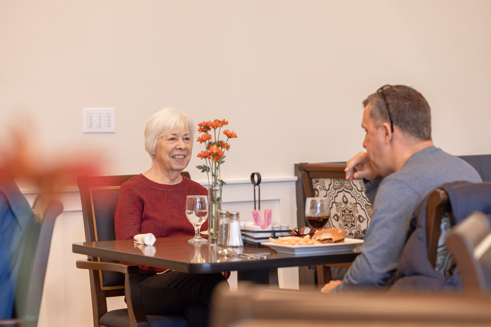 An elderly woman and a middle-aged man sitting at a dining table in a senior living facility, engaged in conversation. The table has a vase with orange flowers, a glass of water, a glass of dark beverage, and a plate with a sandwich. The setting is warm and inviting with cushioned chairs and a neutral wall background.