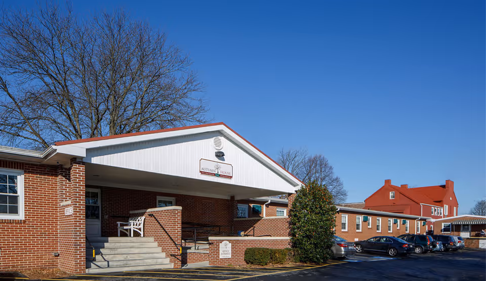 Exterior view of Autumn House East facility showing a brick building with a covered entrance, steps, a white bench, and a parking lot with several cars under a clear blue sky.