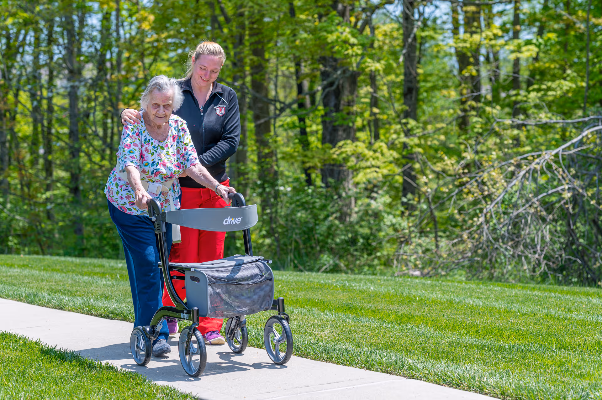 An elderly woman using a walker is being assisted by a caregiver as they walk on a sidewalk surrounded by green grass and trees in an outdoor setting.
