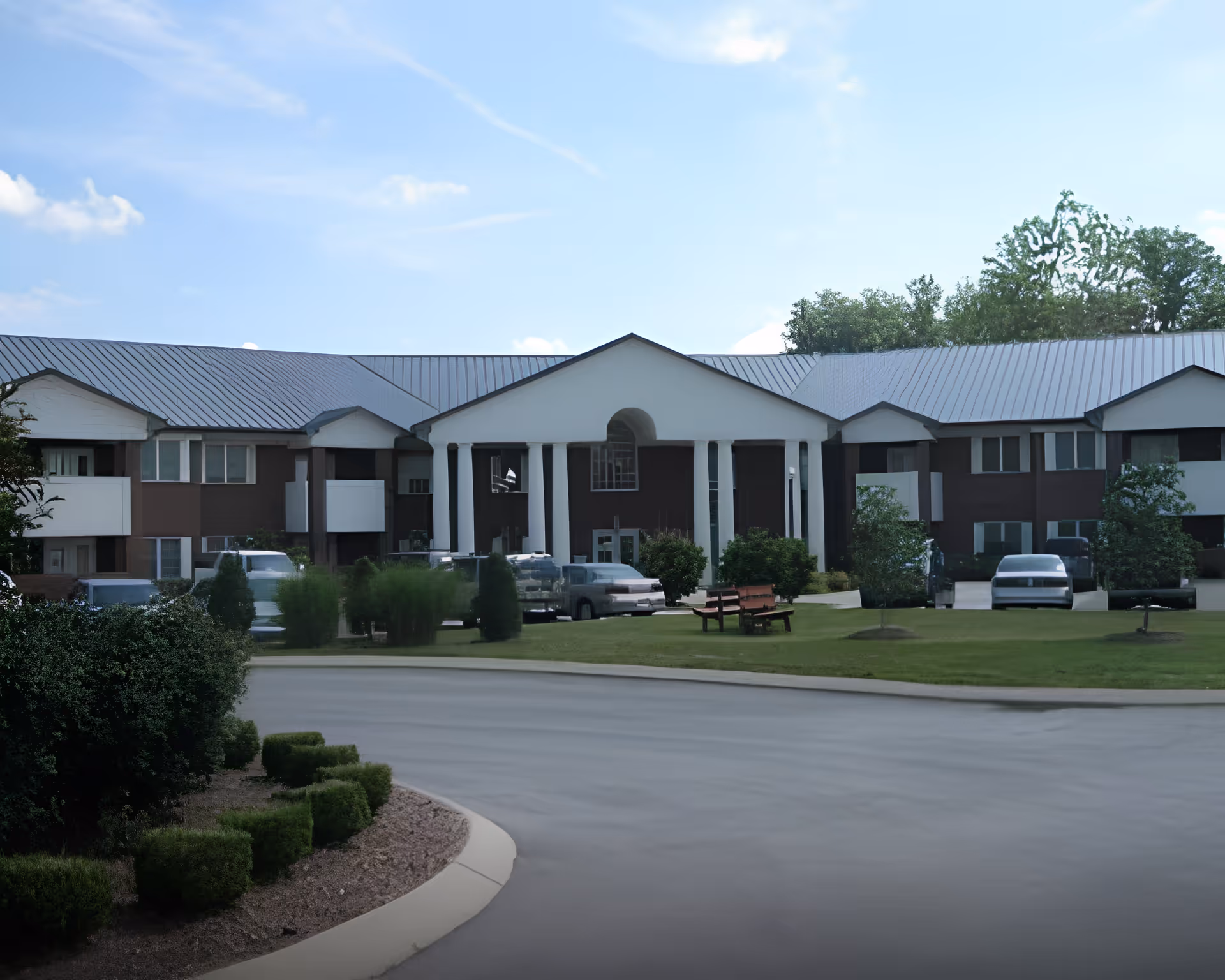 Front exterior view of a senior living facility building with a large white portico supported by columns, surrounded by green lawns, bushes, and parked cars under a blue sky with some clouds.
