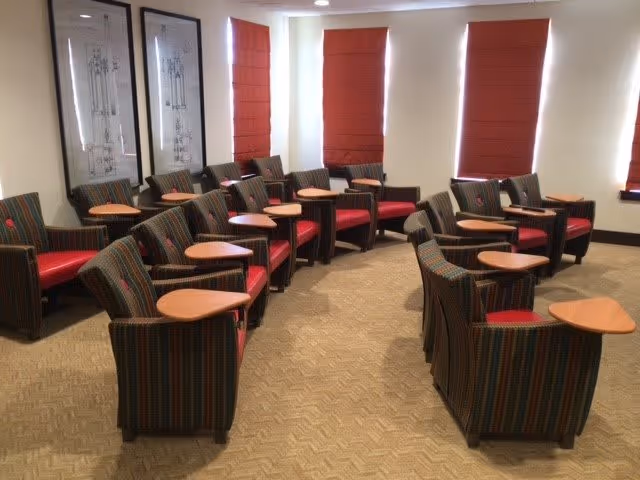 Room with rows of striped armchairs each with a small attached wooden table, arranged under red window shades.