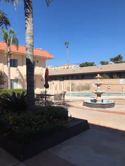 Courtyard with a fountain, patio table and chairs, a fenced swimming pool and a two-story building under a clear blue sky.