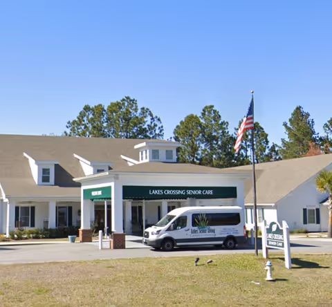 Front exterior of Lakes Crossing Senior Care with a shuttle van parked under the entrance canopy and an American flag in front.