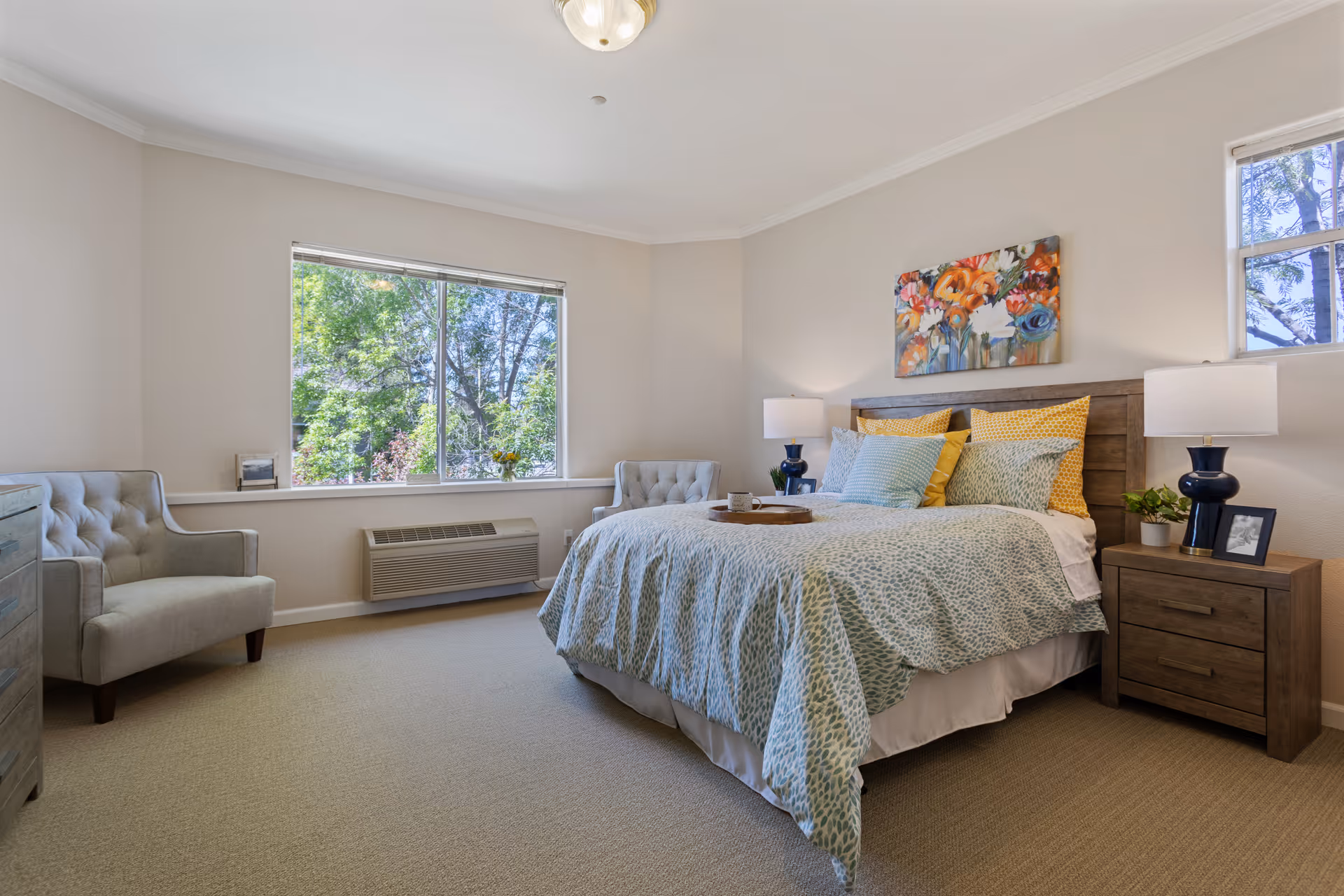 Bright, neatly staged bedroom with a made bed, wooden headboard, bedside tables and lamps, upholstered chairs, and a large window showing trees.