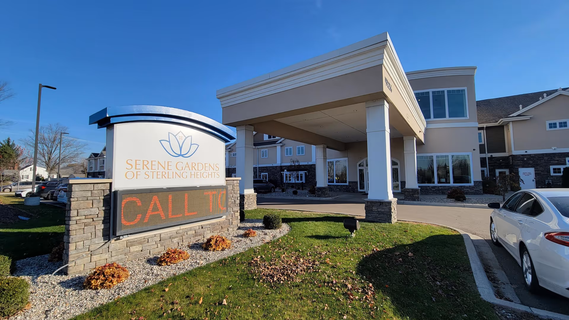 Exterior view of Serene Gardens of Sterling Heights senior living facility showing the main entrance with a covered drop-off area, a stone and brick sign with the facility name and a digital message board, parked cars, and a clear blue sky.