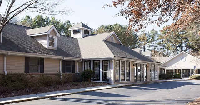 Exterior view of a single-story building with beige siding and multiple windows, surrounded by trees with autumn foliage. The building has a pitched roof with dormer windows and a small cupola. A paved driveway runs alongside the building.