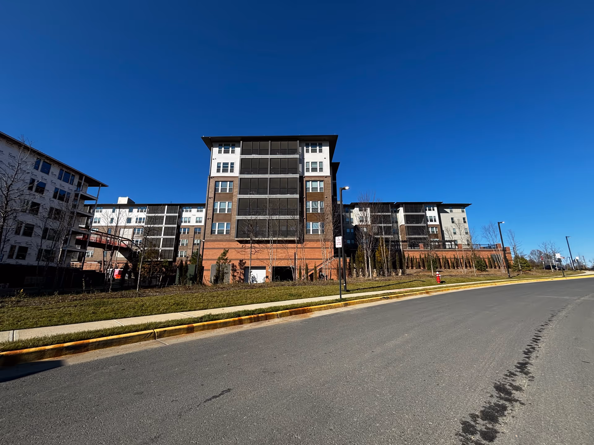 Front exterior view of a multi-story senior living building with brick and glass facade beside a road under a clear blue sky.
