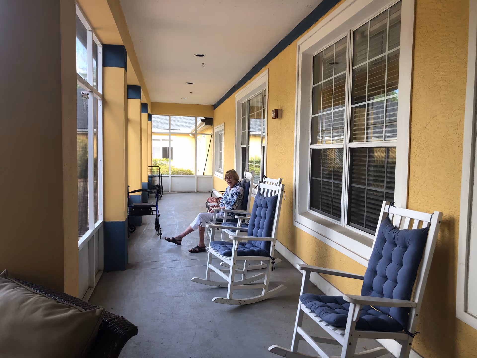 A covered outdoor patio area with yellow walls and white-framed windows. There are several cushioned rocking chairs lined up along the wall. An elderly woman is sitting on one of the chairs, smiling. The patio is enclosed with screened windows allowing natural light in.