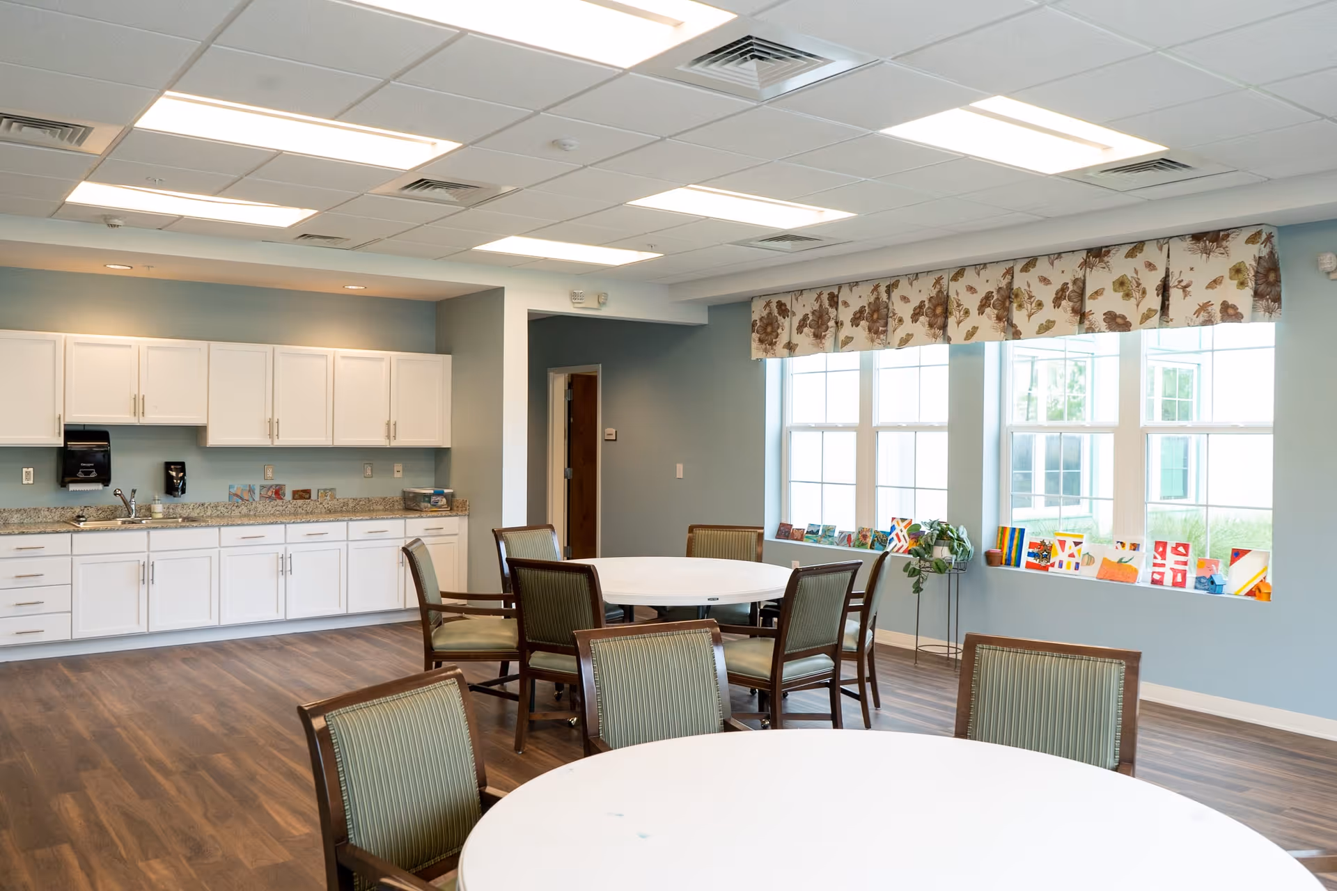 A bright dining area with round white tables and green cushioned chairs. The room features large windows with floral valances, letting in natural light. Along one wall, there is a kitchen area with white cabinets, a granite countertop, a sink, and a paper towel dispenser. The floor is wood-style, and the ceiling has recessed lighting panels.