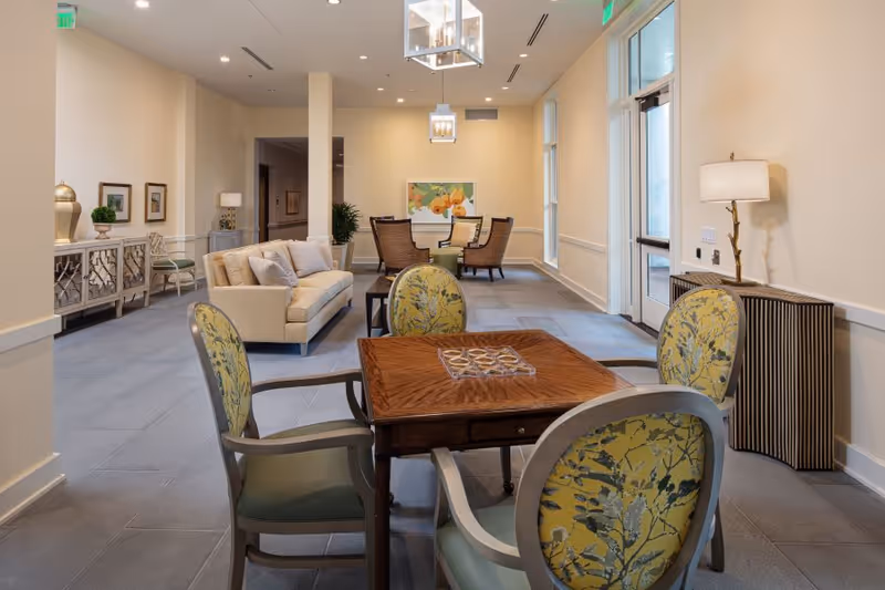 Bright communal lounge with a game table and patterned chairs in the foreground and a sofa and dining area in the background.