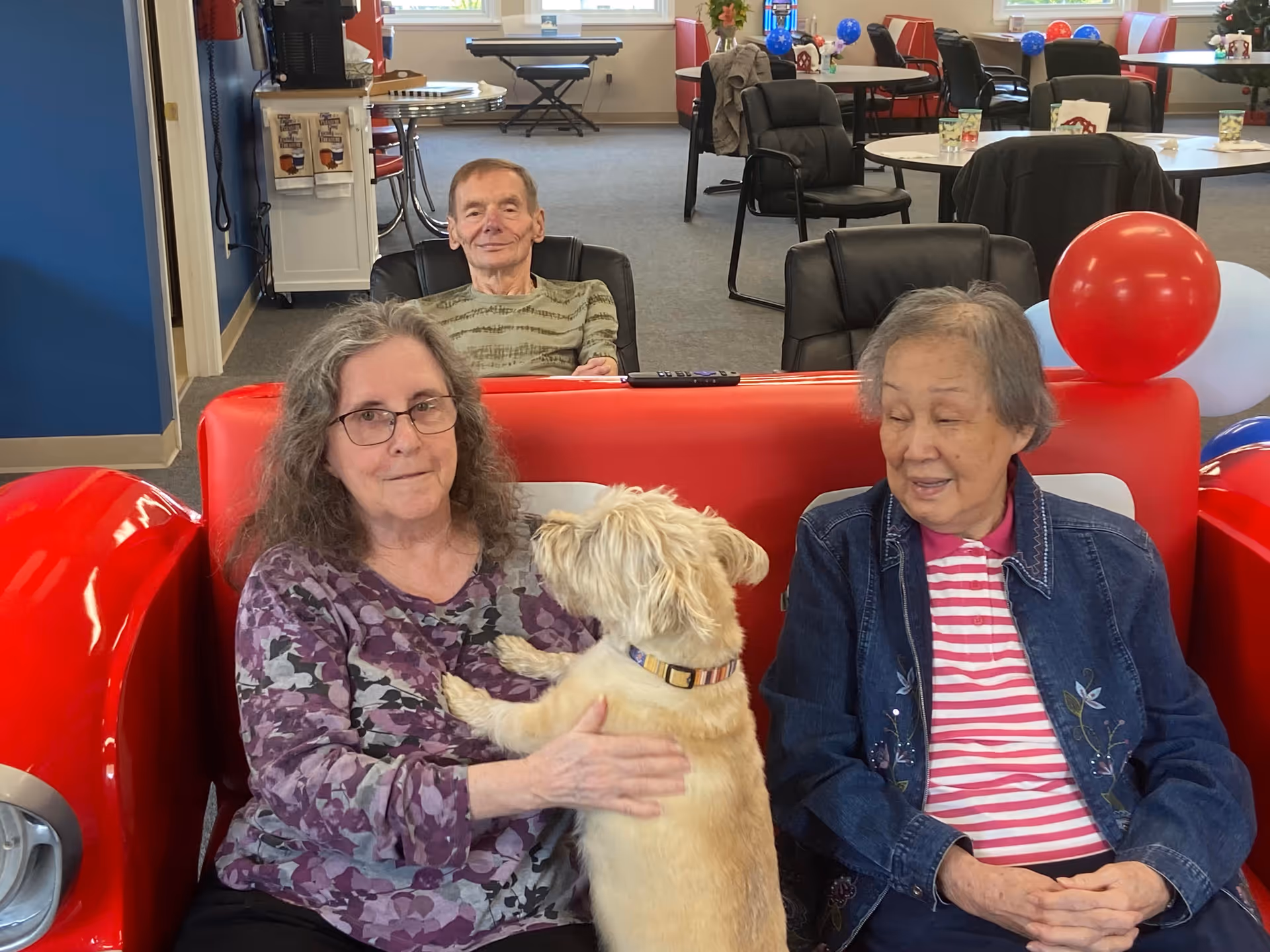 Two elderly women sitting on a red couch in a common area, one woman holding a small light-colored dog. An elderly man is seated behind them in a black chair. The room has tables and chairs, some decorated with red, white, and blue balloons.